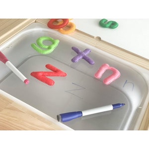 May include: A white dry-erase board with a clear plastic tray underneath. The tray contains felt letters in various colours, including pink, red, green, and purple. Two white dry-erase markers are also visible.