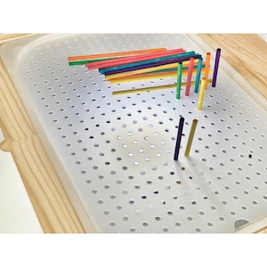 May include: A white pegboard with a grid of holes, featuring colourful wooden dowels arranged in a pattern. The dowels are various shades of red, orange, yellow, green, blue, and purple.