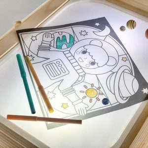 May include: A light-up drawing board with a black and white astronaut, planets, and stars. The board is illuminated from below, and there are three coloured markers on the surface.