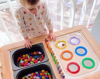 Op de afbeelding: Een houten speeltafel met twee zwarte bakken gevuld met kleurrijke houten vormen. De tafel heeft ook een wit bord met gekleurde cirkels en de woorden "Rood", "Oranje", "Geel", "Groen", "Blauw", "Paars" erop geschreven. Een kind speelt met de speelgoed.