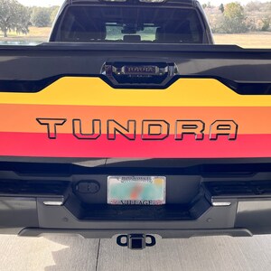 Puede incluir: Vista trasera de una camioneta Toyota Tundra negra con un diseño retro colorido. El diseño presenta rayas horizontales en amarillo, naranja y rojo, con la palabra "TUNDRA" en letras negras. La camioneta tiene una placa y un enganche de remolque.