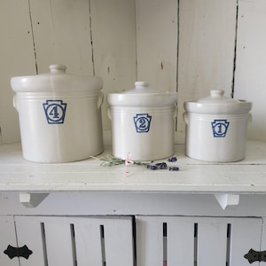 May include: A set of three off-white ceramic canisters with lids, each featuring a blue stamped keystone emblem containing a number. The canisters are labeled with the numbers four, two, and one, arranged in descending order of size from left to right.