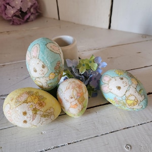 May include: Four Easter eggs decorated with glitter and images of bunnies. The eggs are in shades of blue and yellow.