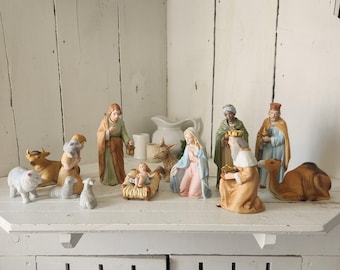 Può includere: Un set presepio in ceramica con statuine della Sacra Famiglia, dei tre re magi, di un pastore, di un asino, di una mucca e di tre pecore. Le figure sono dipinte in tonalità tenui di blu, beige, verde e marrone, con Gesù bambino nella mangiatoia.