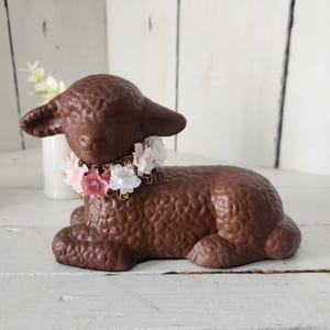 May include: A brown lamb figurine with a textured surface, wearing a floral collar of pink and white flowers. The lamb is lying down on a white surface, with a white vase and flowers in the background.