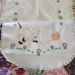 May include: White embroidered table runner featuring a playful kitten with a ball of yarn. The design includes blue floral accents and a delicate lace trim. The cat is light gray with brown paws and ears. The runner is a charming addition to any home decor.