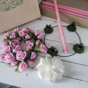 May include: A collection of floral craft supplies, including a pink rose wreath, pink twisted candles, a white satin bow, and green candle holders. A cardboard box with a rose design is also present. The items are arranged on a white wooden surface.