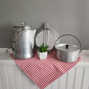 May include: A collection of vintage camping cookware, including a metal kettle, a round mess kit, and a small pot with a handle. A small potted succulent sits on a red and white checkered cloth.