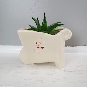 May include: White ceramic planter shaped like a wheelbarrow with red flower accents. A small green succulent plant is growing in the planter.