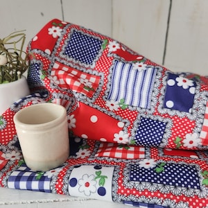 May include: A cream-colored ceramic container sits on a folded red, white, and blue patchwork fabric. The fabric features polka dots, gingham, and floral patterns. The overall theme is a vintage, country style.