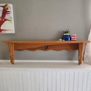 May include: A wall-mounted wooden shelf featuring a scalloped front edge with a central heart-shaped cutout. The shelf has two side supports and a natural wood grain finish. A small blue pot with a green plant and a red and white striped jar sit on top.