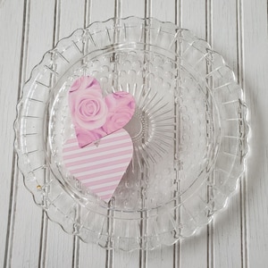 May include: A clear glass cake plate with a scalloped edge and a raised, textured centre. Two pink paper hearts with a floral and striped design are placed on the plate.