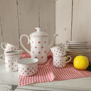 May include: A white porcelain coffee set featuring a red clover-like pattern, including a tall coffee pot with a lid, a creamer, a sugar bowl, four cups, and a stack of saucers. A single yellow lemon sits beside the stack of white saucers.
