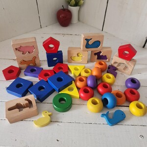 May include: A collection of wooden building blocks and beads in various geometric shapes and animal silhouettes. The set includes red, blue, yellow, green, orange, and purple pieces, some featuring cutouts of a pig, cat, whale, and duck.