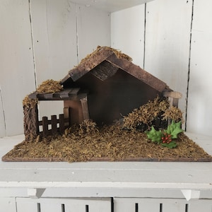 May include: A wooden nativity stable structure featuring a peaked roof, a small picket fence on the left side, and a flat base covered in dried green moss. The dark brown wood is accented with patches of moss and a sprig of green holly with red berries.