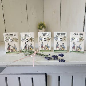 May include: Five rectangular ceramic spice containers labeled Cinnamon, Salt, Pepper, Allspice, and Nutmeg. Each white container features a small knob and a colorful illustration of a person in historical attire. A sprig of dried lavender is tied with a pink ribbon.