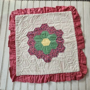 May include: A square, handmade quilt with a floral design. The center features a flower with green, pink, and yellow petals, surrounded by white hexagon shapes. A ruffled, pink border frames the quilt, which is placed on a white, wooden surface.