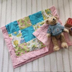 May include: A patchwork baby quilt with a pink border and a teddy bear lying on top. The quilt is made of different floral fabrics in shades of green, blue, and white.