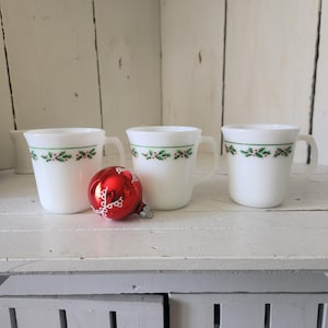 May include: Three white mugs with a green and red holly berry design. A red Christmas ornament with a white lace design sits in front of the mugs. The mugs are on a white shelf.