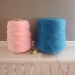 May include: Two large cones of yarn, one in a soft pastel pink and the other in a vibrant bright blue. Both cones feature a fuzzy, textured fiber appearance. A small, delicate white flower with green leaves sits between the two yarn cones.