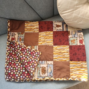 May include: A patchwork quilt featuring a variety of square fabric panels in brown, yellow, and red tones, with patterns including wavy lines, floral motifs, and outdoor themes. The reverse side displays a brown fabric with colorful polka dots.