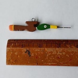 May include: A hand-painted wooden crochet hook featuring a mallard duck head design with a green head, yellow beak, and brown body. The hook measures approximately 3.5 inches in length when placed alongside a wooden ruler for scale.