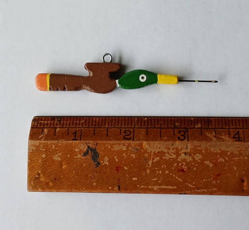 May include: A hand-painted wooden crochet hook featuring a mallard duck head design with a green head, yellow beak, and brown body. The hook measures approximately 3.5 inches in length when placed alongside a wooden ruler for scale.