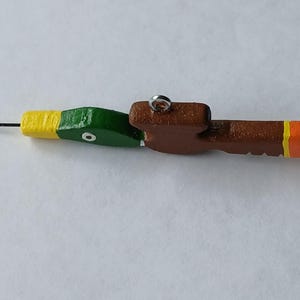 May include: A wooden fishing lure shaped like a duck head and body, painted with a green head, yellow neck, brown body, and orange tail tip. It features a metal eyelet on top for attachment and a thin metal wire extending from the front beak area.