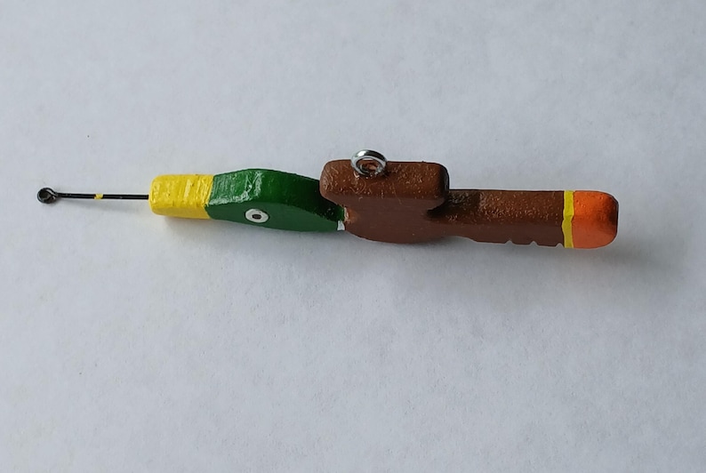 May include: A wooden fishing lure shaped like a duck head and body, painted with a green head, yellow neck, brown body, and orange tail tip. It features a metal eyelet on top for attachment and a thin metal wire extending from the front beak area.