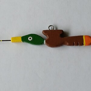 May include: A crochet hook with a handle shaped like a mallard duck. The duck head is green with a yellow beak and white eye detail. The body is brown with a bright orange tip and a small metal loop attached to the top for hanging or storage.