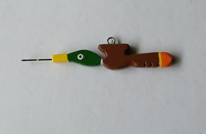 May include: A crochet hook with a handle shaped like a mallard duck. The duck head is green with a yellow beak and white eye detail. The body is brown with a bright orange tip and a small metal loop attached to the top for hanging or storage.