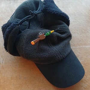 May include: A dark olive green fleece cap with black ear flaps and a black brim. A small, brown and green duck-shaped pin with a yellow beak and a silver metal eyelet is attached to the front of the cap, featuring a thin, needle-like protrusion.