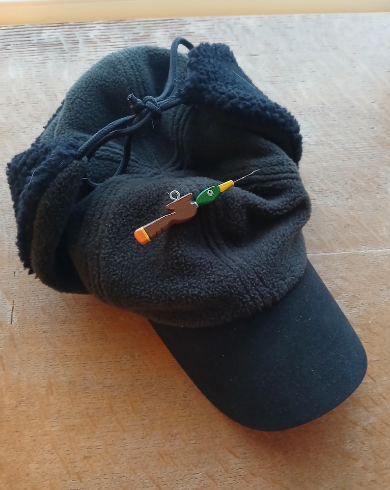 May include: A dark olive green fleece cap with black ear flaps and a black brim. A small, brown and green duck-shaped pin with a yellow beak and a silver metal eyelet is attached to the front of the cap, featuring a thin, needle-like protrusion.
