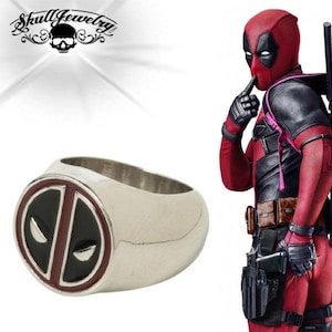 Puede incluir: Un anillo de plata con un logotipo de Deadpool rojo y negro. El anillo es de estilo sello con un logotipo en relieve. El logotipo es una versión estilizada de la máscara de Deadpool.
