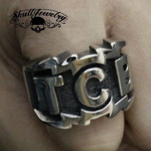 Puede incluir: Un anillo de plata con las letras "FCB" en relieve. El anillo se lleva en el dedo de una persona.