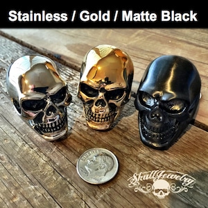 May include: Three skull rings in different finishes: stainless steel, gold, and matte black. The rings are all the same design, with a detailed skull face. A 25 pence coin is included for size reference.