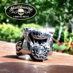 May include: A silver-colored ring with a detailed motorcycle engine design. The ring is set against a blurred green background. The image includes a black oval logo with the text "Skull Jewelry" and a phone number.