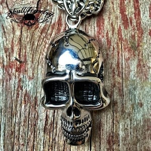 Puede incluir: Un colgante de calavera de plata con costuras detalladas en la cabeza y una cara texturizada. El colgante cuelga de una cadena de plata.