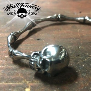 May include: A silver skull bracelet with a bone design. The bracelet is open and has a skull charm on the end.