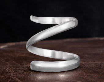 Könnte beinhalten: Ein silberfarbenes, spiralförmiges Armband. Das Armband besteht aus glattem, glänzendem Metall und hat ein minimalistisches Design. Das Armband wird auf einer braunen, strukturierten Oberfläche mit dunklem Hintergrund gezeigt.