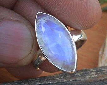 Könnte beinhalten: Ein Silberring mit einem großen, ovalen Mondstein. Der Mondstein hat eine perlmuttartige, blau-weiße Farbe und ist in einem schlichten Silberband gefasst. Der Ring wird vor einem natürlichen Holzuntergrund gezeigt.
