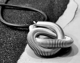 Puede incluir: Un collar de metal plateado con diseño de resorte en espiral. El collar está sobre una superficie blanca y negra.