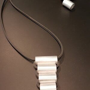 May include: A silver necklace with a layered pendant made of stacked silver rings. The necklace is on a black cord.