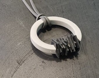 Puede incluir: Un collar gris con un colgante circular blanco y un diseño geométrico negro. El collar está hecho de cuero y tiene un estilo simple y moderno.