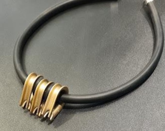 May include: A black rubber choker necklace with a gold-colored metal accent. The metal accent features a series of curved, stacked elements. The necklace has silver-colored clasps at each end.