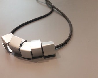 Puede incluir: Un collar plateado con un diseño geométrico. El collar está hecho de varios cuadrados plateados que están conectados entre sí. El collar está en un cordón negro.