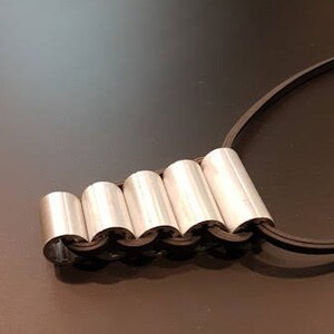 May include: A silver necklace with a chain made of linked cylinders. The necklace is on a black cord.