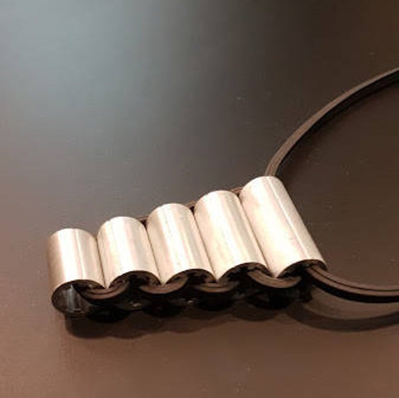 May include: A silver necklace with a chain made of linked cylinders. The necklace is on a black cord.