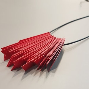 Może przedstawiać: Czerwony naszyjnik z papieru o kształcie wachlarza. Naszyjnik wykonany jest z wielu małych, złożonych kawałków papieru.