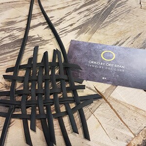 May include: Black leather necklace with a woven design. The necklace is on a wooden surface. The card reads "Oriko by Ort Efan Jewelry Designer".